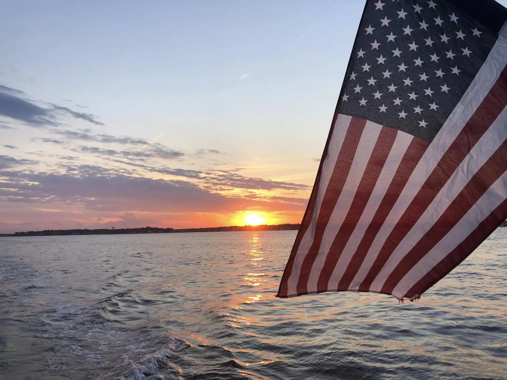 American flag with red and white stripes and a blue field of stars, waving above water at sunset.